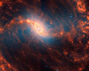 Image captured by the James Webb Telescope showcasing the NGC 1365 spiral galaxy. Vivid hues of orange and blue dominate the scene, highlighting the intricate swirls and spirals of stardust and gas. The galaxy's bright core emits a yellowish glow, with numerous stars dotting the cosmic landscape. Darker regions within the spirals suggest areas dense with cosmic material, obscuring the light behind them.